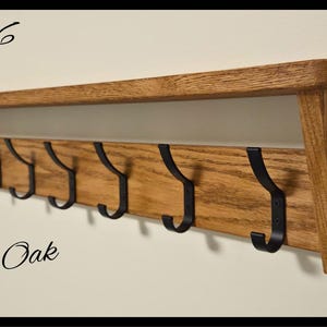 May include: A wall-mounted golden oak shelf featuring seven black metal hooks attached to a horizontal wooden board. The text Code 06 is visible in the top left, Golden Oak in the bottom left, and a circular logo for Nilsa Prairie in the bottom right.