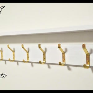 May include: A white wooden wall-mounted coat rack featuring a top shelf and seven gold-toned metal hooks. The text Code 09 is in the top left corner, the word White is in the bottom left, and a circular NILSA PRAIRIE logo is in the top right.