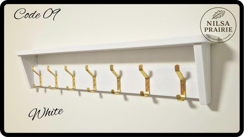 May include: A white wooden wall-mounted coat rack featuring a top shelf and seven gold-toned metal hooks. The text Code 09 is in the top left corner, the word White is in the bottom left, and a circular NILSA PRAIRIE logo is in the top right.