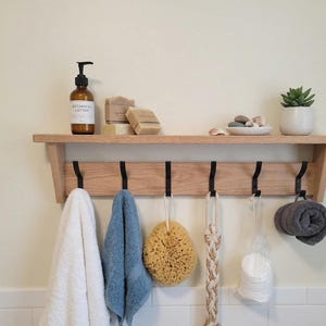 May include: A light-toned wooden wall shelf featuring six black metal hooks. The shelf holds a brown bottle of botanical lotion, two rectangular soap bars, a small white bowl with stones, and a potted succulent. Hanging items include towels, a sponge, and a washcloth.