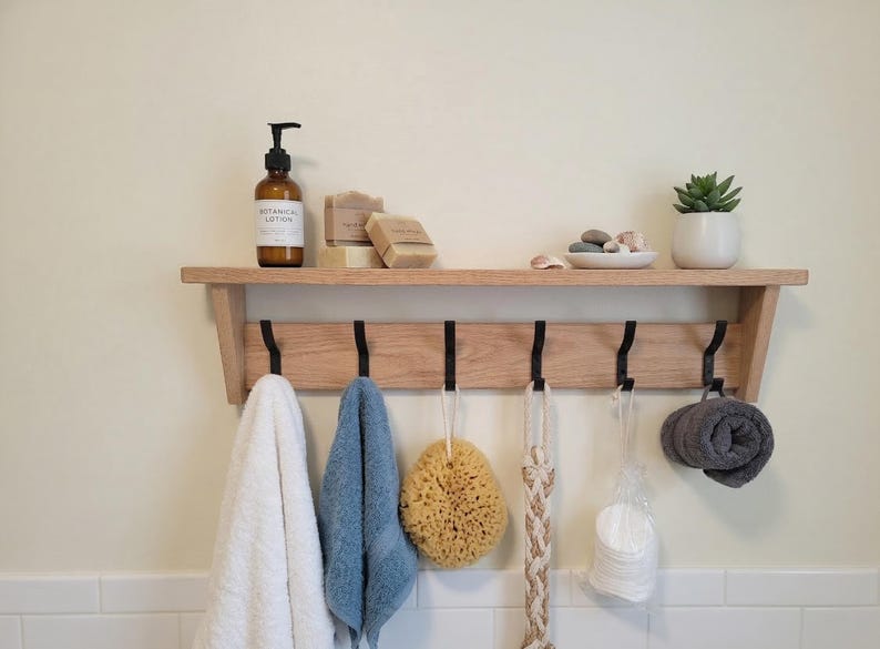 May include: A light-toned wooden wall shelf featuring six black metal hooks. The shelf holds a brown bottle of botanical lotion, two rectangular soap bars, a small white bowl with stones, and a potted succulent. Hanging items include towels, a sponge, and a washcloth.