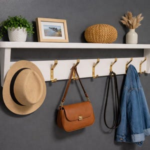 May include: A white wooden wall-mounted shelf featuring a top display ledge and a lower rack with six gold-toned metal hooks. The shelf holds a potted green plant, a framed photograph, a woven basket, and a white vase with dried floral stems.