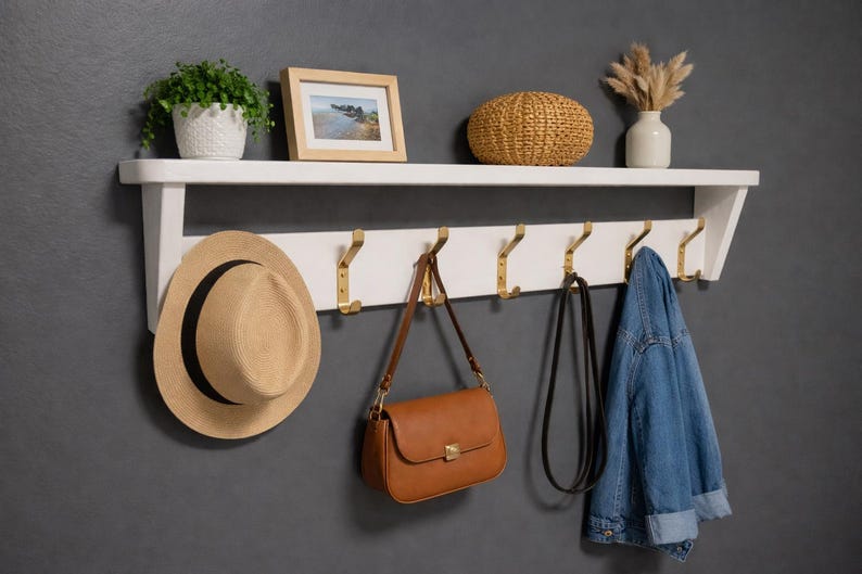 May include: A white wooden wall-mounted shelf featuring a top display ledge and a lower rack with six gold-toned metal hooks. The shelf holds a potted green plant, a framed photograph, a woven basket, and a white vase with dried floral stems.