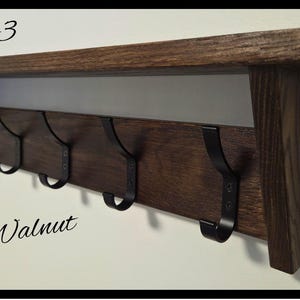 May include: A dark walnut wood wall shelf featuring five black metal hooks attached to the front panel. The text Code 03 is printed in the top left corner, and a circular logo with the text NILSA PRAIRIE HANDCRAFTED IN CANADA is in the bottom right.