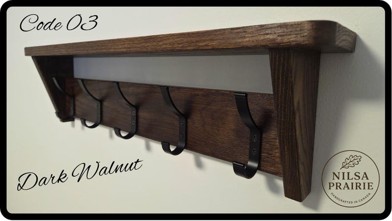 May include: A dark walnut wood wall shelf featuring five black metal hooks attached to the front panel. The text Code 03 is printed in the top left corner, and a circular logo with the text NILSA PRAIRIE HANDCRAFTED IN CANADA is in the bottom right.