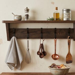 May include: A dark brown wooden wall shelf with five black metal hooks. Hanging from the hooks are a grey and white striped kitchen towel, a metal whisk, a set of wooden measuring spoons, a copper ladle, and a wooden spatula.