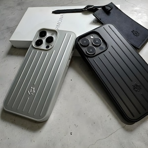 Op de afbeelding: Twee telefoonhoesjes, één lichtgrijs en één zwart, met een geribbeld ontwerp en een logo. Een witte tas met het woord "RIMOWA" staat op de achtergrond, samen met een zwart leren label.