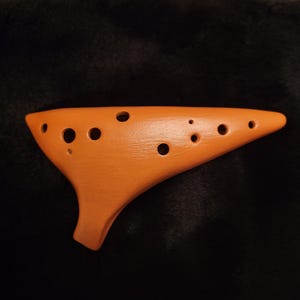 May include: An orange ocarina, a wind instrument, is displayed. The instrument has a teardrop shape with multiple finger holes. The ocarina is made of a solid material and has a smooth, matte finish. The instrument is approximately 6 inches long.