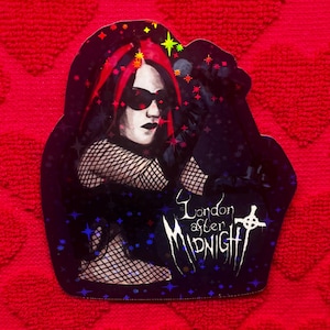 May include: A sticker featuring a gothic illustration of a person with red hair and sunglasses. The person is wearing black clothing and fishnet. The sticker has the text "London After Midnight" with a cross symbol.