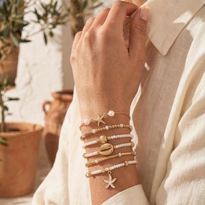 Könnte beinhalten: Ein Set aus fünf geschichteten Armbändern mit weißen Perlen, goldfarbenen Akzenten und nautischen Anhängern. Die Kollektion umfasst einen goldenen Seestern, eine mittige Kaurischnecke und einen hängenden weißen Seestern-Anhänger.