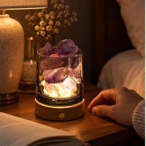 May include: A cylindrical glass vessel filled with raw, purple amethyst crystals resting on a circular light-colored wooden base. The base features a small, round metallic button on the front, and the crystals are illuminated from below by a warm light.