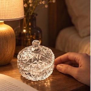 May include: A clear glass trinket dish with a faceted geometric pattern and a matching lid featuring a small knob handle. The circular container has a textured surface that catches the light, creating a sparkling effect on the wooden surface.