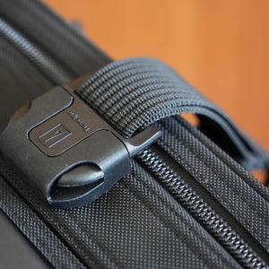 May include: A black luggage strap with a plastic buckle featuring the Travelpro logo. The strap is attached to a black textured suitcase with a visible zipper track. The buckle mechanism is secured over the fabric surface of the travel bag.