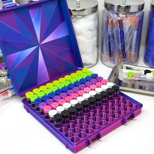 May include: A purple and blue iridescent storage case with a geometric star pattern on the lid. The interior features a grid of circular slots holding small glass vials with colorful caps in neon green, blue, pink, white, and black.