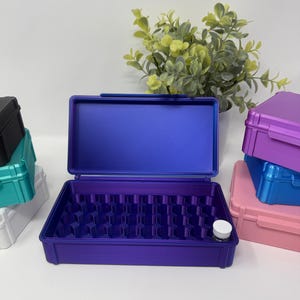 May include: A rectangular purple storage case with a hinged lid and an interior grid pattern designed for small items. The case is open, revealing a deep purple base with a single small white-capped vial placed in one of the grid compartments.