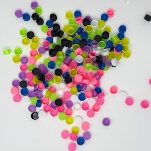 May include: A large collection of small, circular plastic caps in a variety of vibrant colors including neon pink, lime green, bright blue, purple, black, and white. The caps are scattered across the frame, showing their hollow interiors.