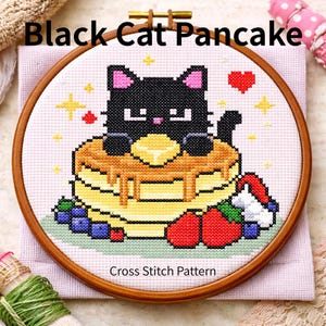 以下が含まれることがあります： 木製のフレームに入ったクロスステッチ刺繍。黒猫がバターとシロップのかかったパンケーキの上にいます。「Black Cat Pancake」と「Cross Stitch Pattern」の文字と、ブルーベリーとイチゴのデザインです。