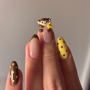 Könnte beinhalten: Eine Hand mit gepflegten Nägeln. Ein Nagel zeigt einen braunen Affen, der eine gelbe Banane hält. Andere Nägel haben braune und weiße Tupfen und eine braune French-Spitze. Die Nägel sind in Braun-, Gelb- und Nude-Tönen lackiert. Nagelkunst.