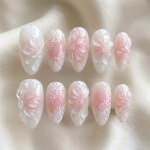 Könnte beinhalten: Ein Set aus zehn künstlichen Nägeln mit floralem Design. Die Nägel zeigen eine Kombination aus Weiß und Rosa mit aufwendigen 3D-Blumen- und Spitzenmustern. Die oval geformten Nägel sind auf einer weichen, weißen Oberfläche angeordnet.