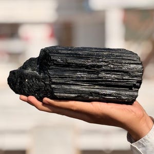 以下が含まれることがあります： 手で持たれた、大きな黒トルマリンの原石。石は深い黒色で、縞模様のテクスチャを持っています。背景はぼやけており、屋外の環境を示唆しています。
