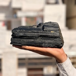 以下が含まれることがあります： 手で持たれた、大きな生の黒トルマリンの結晶。結晶は、暗く不透明な色で、層状で質感のある外観をしています。背景はぼやけており、屋外の環境を示唆しています。