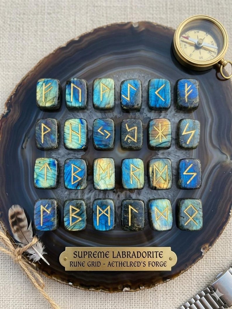 Puede incluir: Un conjunto de veinticuatro piedras de runas de labradorita con s&iacute;mbolos dorados, dispuestas sobre una rebanada de &aacute;gata azul oscuro y gris. Tambi&eacute;n hay una br&uacute;jula dorada y una placa de lat&oacute;n.