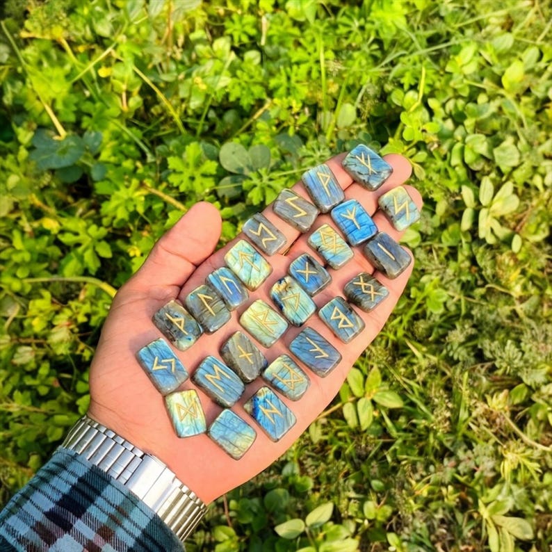 Puede incluir: Colecci&oacute;n de piedras r&uacute;nicas rectangulares azules y verdes con s&iacute;mbolos dorados. Las piedras se sostienen en una mano sobre un fondo de follaje verde. Cada piedra mide aproximadamente 2,5 cm de largo.