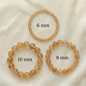 Op de afbeelding: Drie cirkelvormige armbanden gemaakt van doorschijnende goudbruine kralen. De bovenste armband is gelabeld met 6 mm, de onderste rechterarmband met 8 mm en de onderste linkerarmband met 10 mm, wat de grootte van de individuele kralen aangeeft.