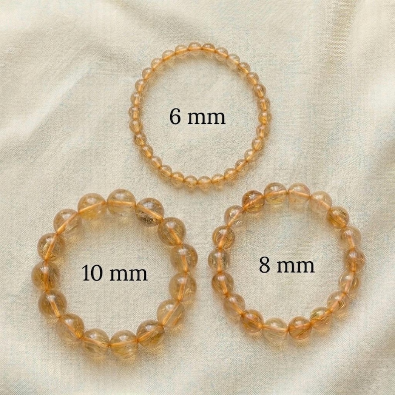 Op de afbeelding: Drie cirkelvormige armbanden gemaakt van doorschijnende goudbruine kralen. De bovenste armband is gelabeld met 6 mm, de onderste rechterarmband met 8 mm en de onderste linkerarmband met 10 mm, wat de grootte van de individuele kralen aangeeft.