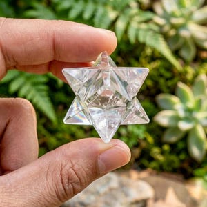 May include: A clear crystal Merkaba star, held between fingers, with multiple facets. The star is transparent, reflecting light. The background features green foliage and succulents, creating a natural setting.