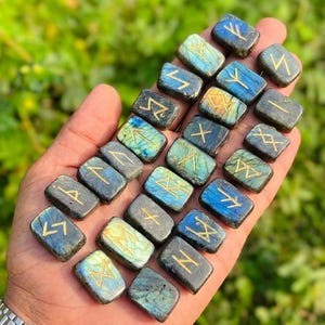 Puede incluir: Una colecci&oacute;n de piedras r&uacute;nicas rectangulares de labradorita, cada una grabada con un s&iacute;mbolo dorado. Las piedras muestran una gama de colores, incluyendo azules, verdes y grises. Las runas se sostienen en una mano, con un fondo verde borroso.