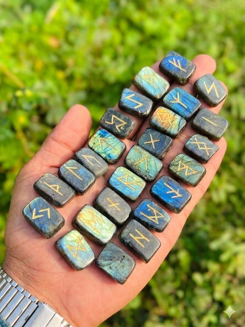 Puede incluir: Una colecci&oacute;n de piedras r&uacute;nicas rectangulares de labradorita, cada una grabada con un s&iacute;mbolo dorado. Las piedras muestran una gama de colores, incluyendo azules, verdes y grises. Las runas se sostienen en una mano, con un fondo verde borroso.
