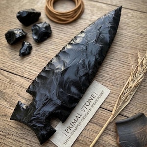 May include: A hand-knapped obsidian spear point with a textured surface, displayed with several black obsidian stones and a bundle of brown cord. The spear point is accompanied by a tag that reads "Primal Stone".