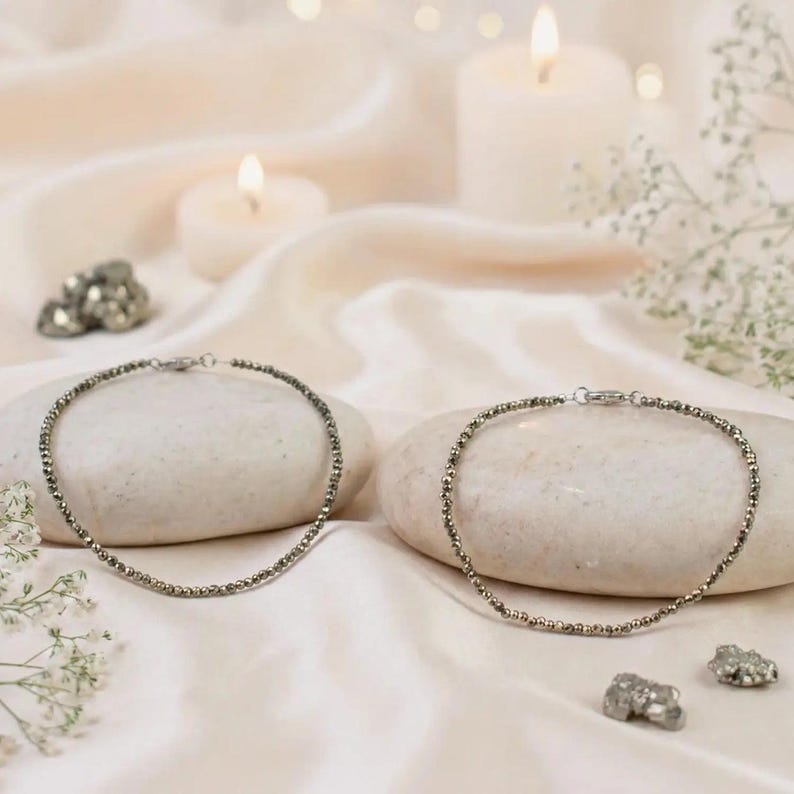 May include: Two delicate beaded anklets with a silver clasp rest on smooth, white stones. The anklets are made of small, dark grey beads. The background features soft, white fabric, candles, and small white flowers, creating a serene aesthetic.