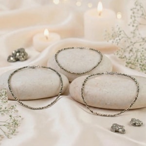 May include: Three delicate beaded necklaces with silver clasps rest on white stones, set against a soft, cream-coloured fabric. The necklaces are composed of small, metallic beads. The scene is illuminated by soft candlelight and sprigs of white flowers.