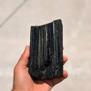 以下が含まれることがあります： 手で持たれた、生の黒トルマリンの結晶。 結晶は、垂直の縞模様のある、粗くテクスチャ加工された表面を持っています。 石は濃い黒色で、光を反射しています。