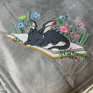 Könnte beinhalten: Eine graue Plüschdecke mit einem gestickten Design. Das Design zeigt einen schlafenden schwarzen Drachen auf einem offenen Buch, umgeben von bunten Blumen. Die Decke hat eine weiche, samtige Textur.