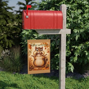 Puede incluir: Una bandera de jardín rectangular que muestra una ardilla de dibujos animados con mejillas llenas y brazos extendidos, rodeada de hojas de otoño y bellotas. El texto "What acorns?" aparece en la parte superior. Cuelga de un poste.