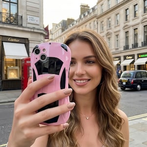 May include: Pink and black car-shaped phone case. The case has a sleek design with a car silhouette. The image is taken outdoors, with a street and buildings in the background. The phone case is held up in front of a person.