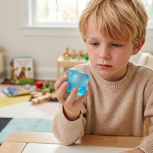 Può includere: Un bambino tiene in mano un giocattolo blu traslucido con la scritta "SEE JOE" stampata sopra. Il giocattolo è tenuto davanti al viso del bambino. Lo sfondo comprende una finestra e un tavolo.
