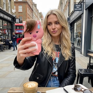 Puede incluir: Una funda de teléfono rosa con forma de oso de dibujos animados con una expresión preocupada. La funda se sostiene frente a una persona con una chaqueta de cuero negra, sentada en una mesa de café al aire libre. Una taza de café y gafas de sol están sobre la mesa.
