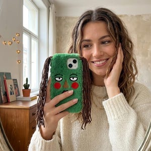 Puede incluir: Una funda de teléfono verde y difusa con un diseño de cara de dibujos animados, con mechones marrones parecidos al cabello. La funda tiene ojos negros y detalles de mejillas rojas. La funda del teléfono se sostiene frente a un espejo.