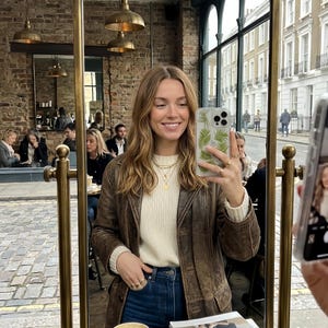 Può includere: Una persona con una giacca di pelle marrone e un maglione color crema si fa un selfie allo specchio. Indossa collane e anelli d'oro. Lo specchio riflette un caffè e una strada.