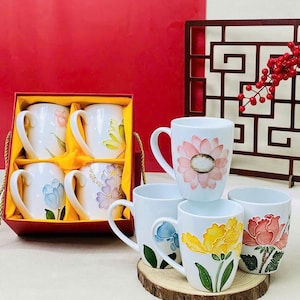 May include: White ceramic mugs featuring floral patterns in pink, yellow, and blue. Some mugs are presented in a red gift box with a gold handle. The mugs are displayed on a wooden coaster.