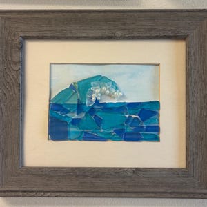 May include: A framed artwork depicting a wave made of blue and turquoise glass pieces. The wave crest features white pearls, set against a light blue background. The artwork is encased in a gray wooden frame.