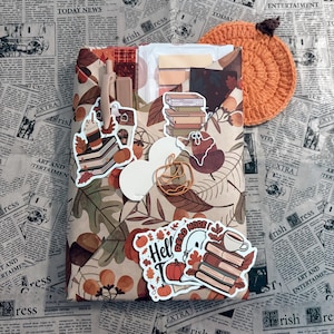 May include: A rectangular package wrapped in paper with a brown and tan autumn leaf pattern, decorated with various stickers of books, pumpkins, and ghosts. A beige pen, a copper pumpkin-shaped paperclip, and a crocheted orange pumpkin are included.