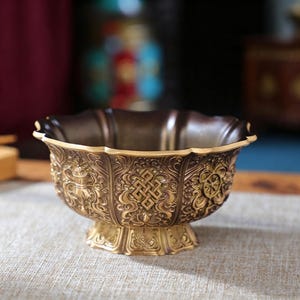 Puede incluir: Un cuenco metálico con acabado en bronce oscuro y reflejos dorados, que presenta un borde festoneado y una base de pedestal. El exterior está decorado con intrincados patrones en relieve, incluyendo un símbolo central de nudo eterno y motivos florales.