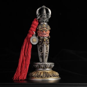 May include: A silver and bronze ritual dagger featuring a detailed wrathful deity face with a red finish, a dragon-engraved blade, and a crown-like pommel. A bright red braided tassel with a circular metal charm hangs from the handle base.