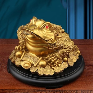 May include: A gold-colored three-legged toad figurine with red gemstone eyes, sitting atop a pile of circular coins and gold ingots. The toad holds a coin in its mouth and features a scroll with the Chinese characters 招財進寶 on its front.
