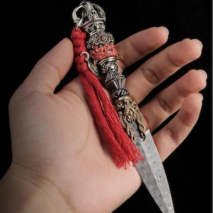 Puede incluir: Una daga ritual phurba con hoja de acero de Damasco con patrón ondulado, mango adornado con una talla de deidad de cara roja y pomo metálico. Una borla larga de cordón trenzado de color rojo brillante está unida a la parte superior.